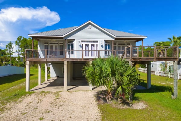 Elevated white cottage with metal roof, wraparound decks, and palm trees in front yard under blue sky