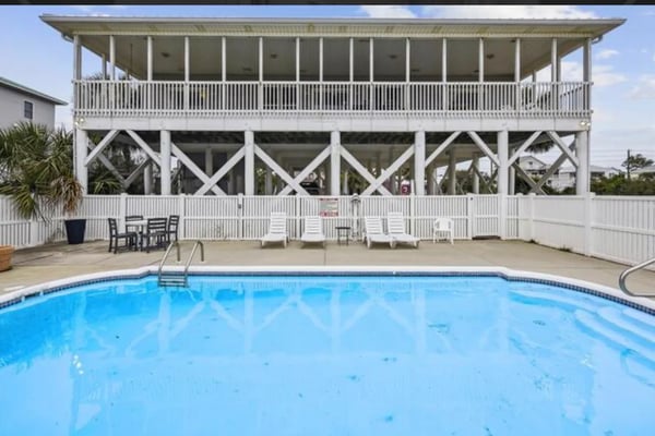 Elevated white beach house with pool, lounge chairs, and covered deck overlooking ocean.